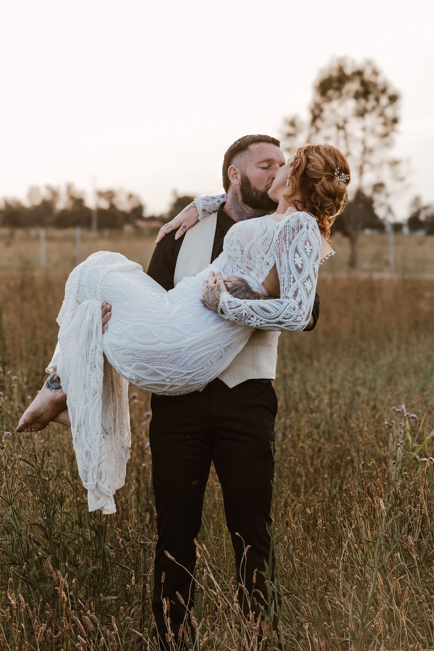 Groom carrying bride in lace dress while kissing in golden field at sunset during outdoor wedding.