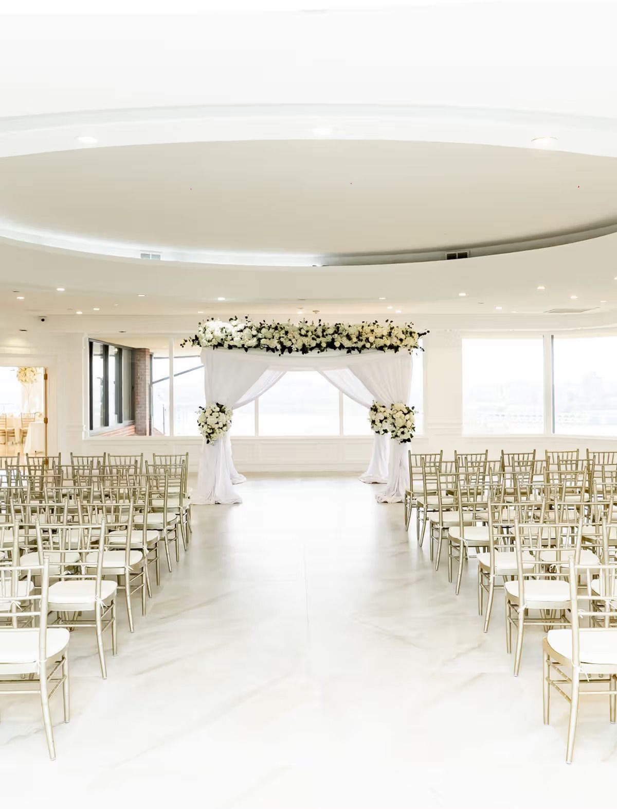 A modern all-white wedding ceremony space with chiavari chairs, floral arch, and floor-to-ceiling windows.