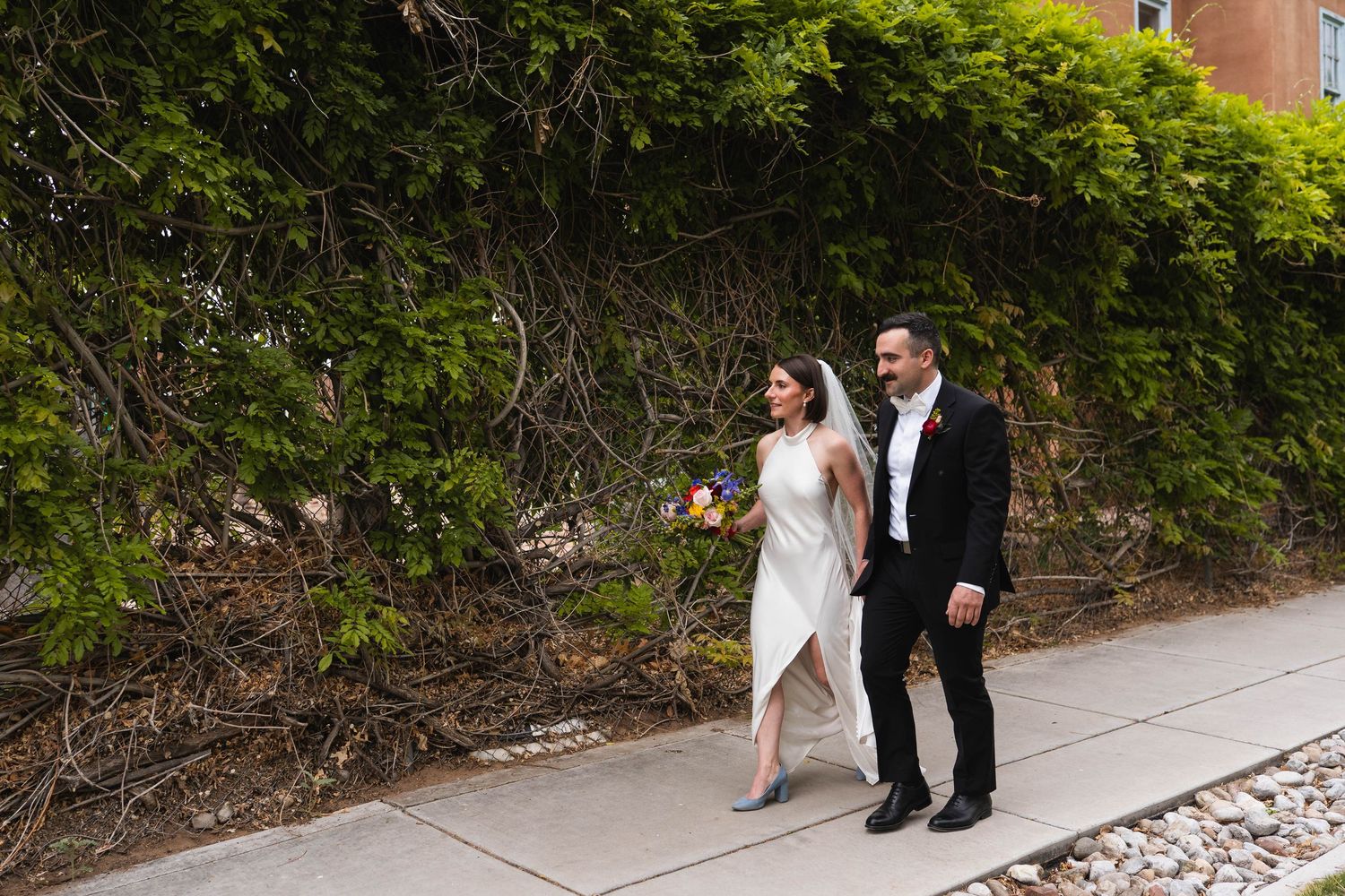 Bride and groom walking hand in hand on a sidewalk beside a lush green ivy wall on their wedding day.