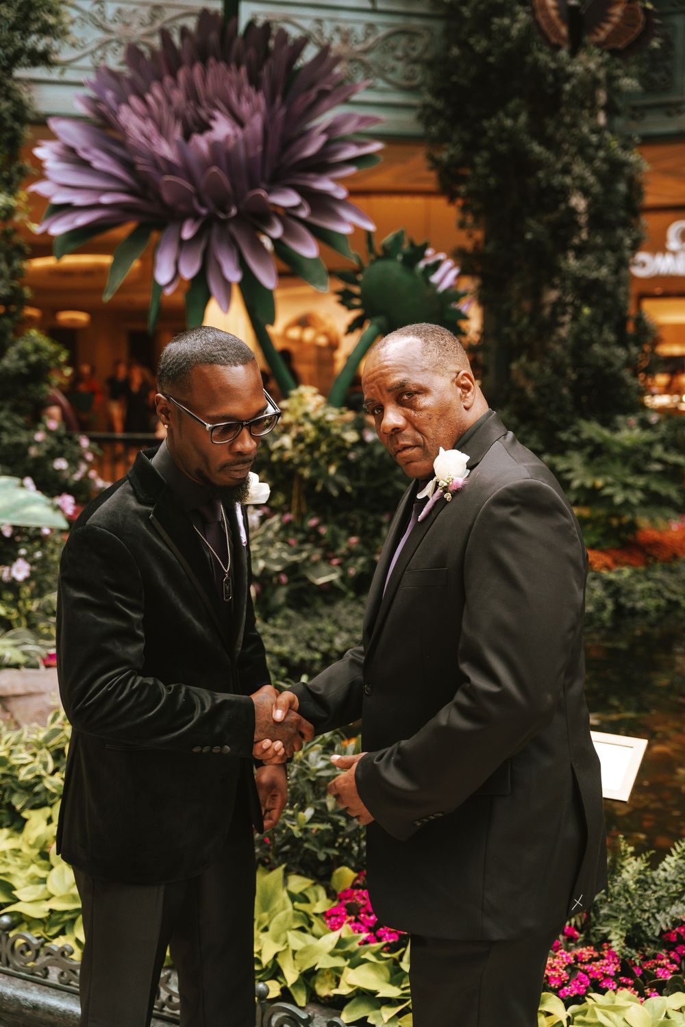 Two men in black suits shaking hands in a garden atrium with colorful flowers and greenery.