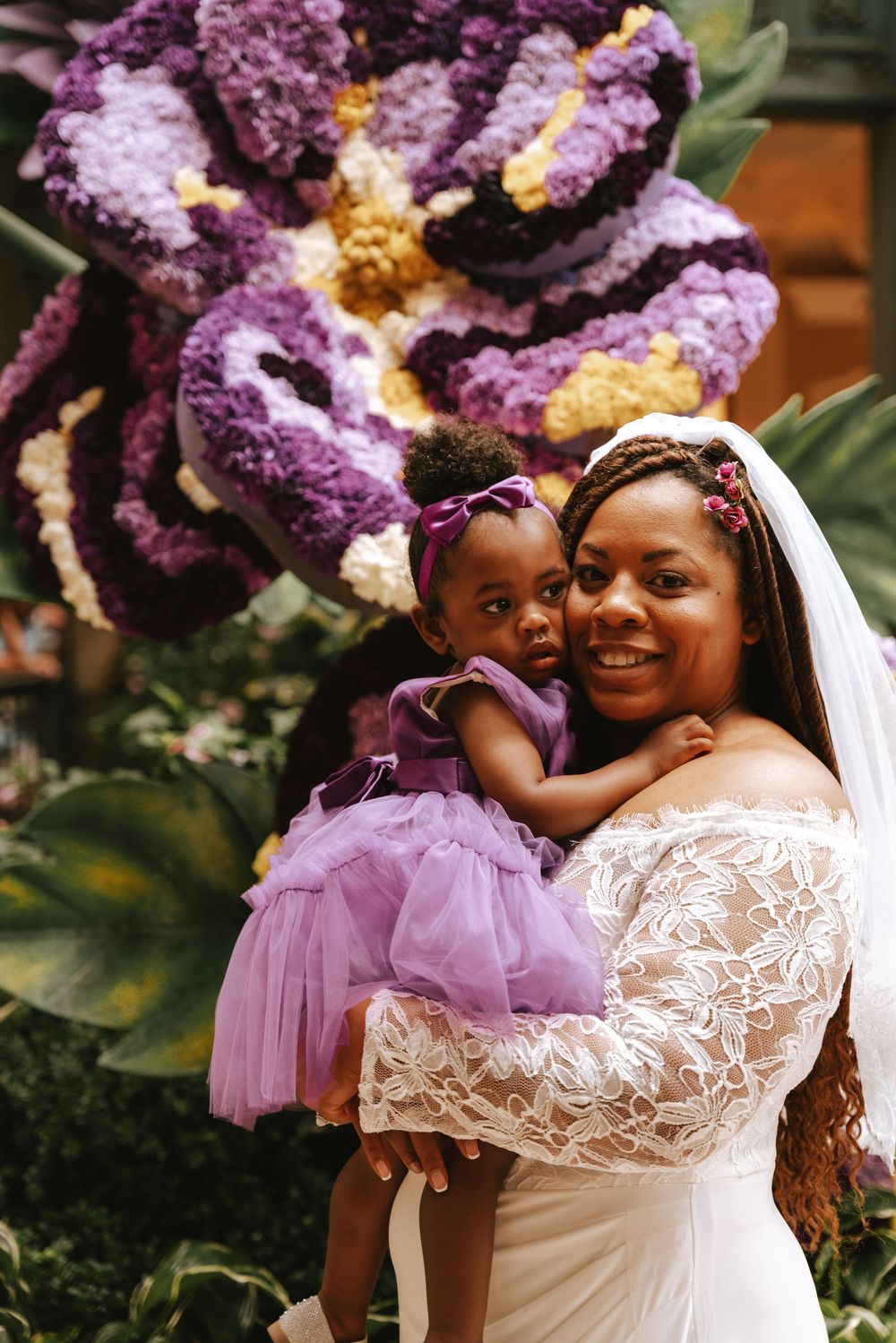 Bride in white lace gown holds young girl in purple tutu dress before large floral display.