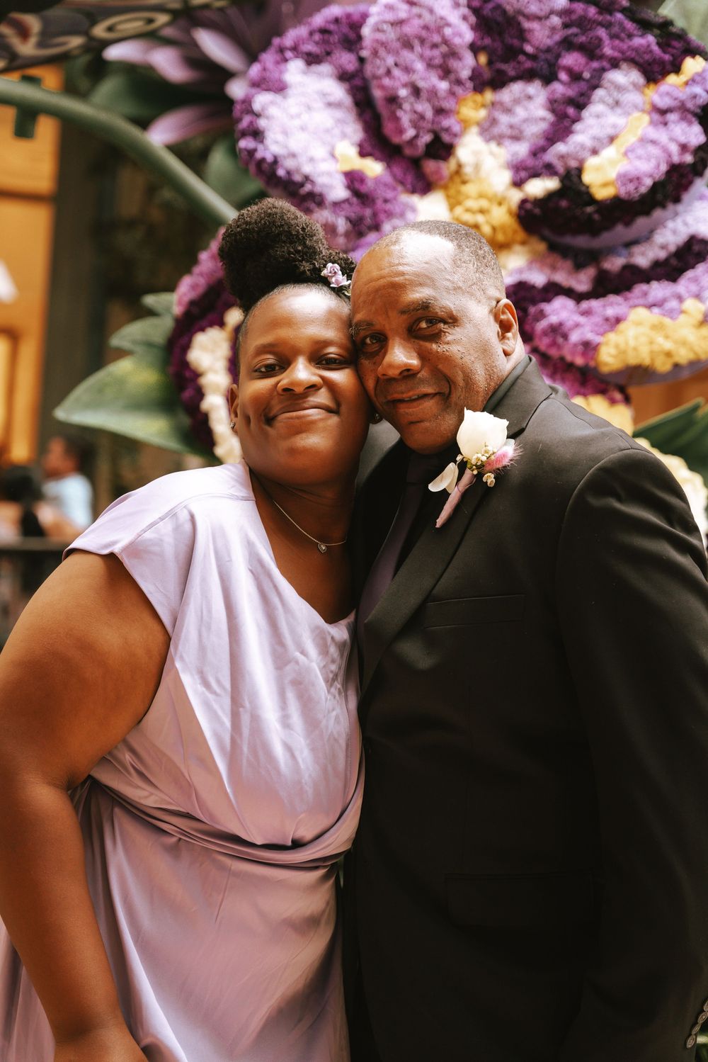 A couple poses together at a wedding, man in black suit with boutonniere, woman in lavender dress, purple floral backdrop.