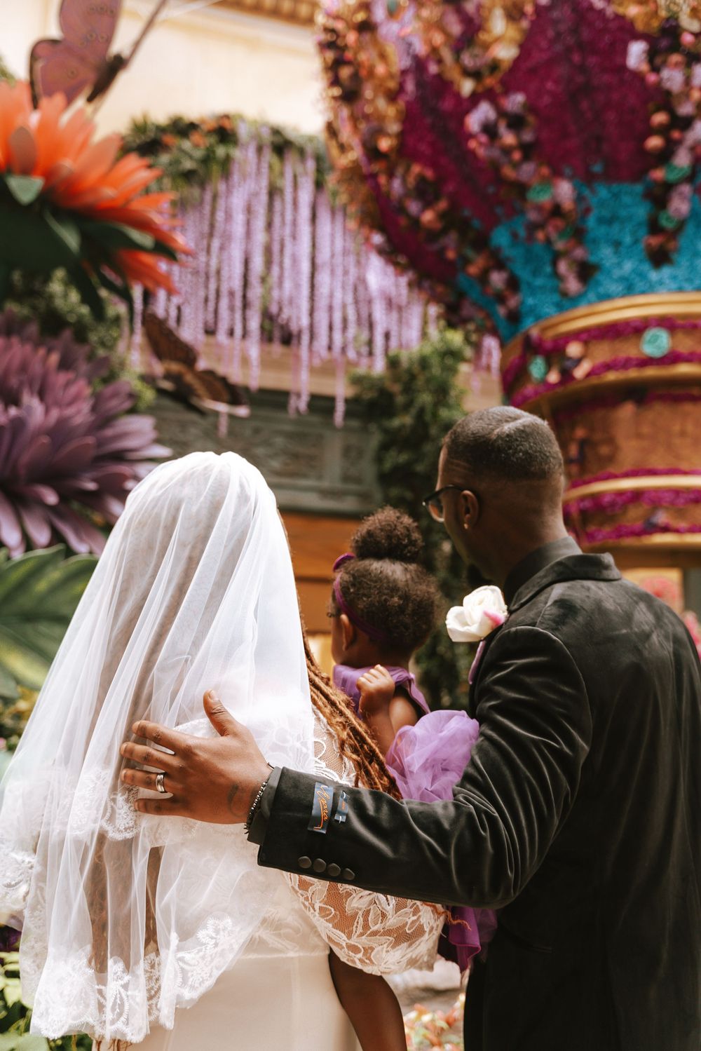 Bride and groom stand at floral wedding altar, bride in lace gown with veil, groom in black suit with boutonniere.