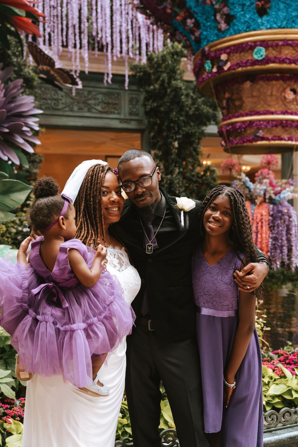 A joyful family poses together at a wedding, with the bride in white, groom, and two daughters in purple attire.