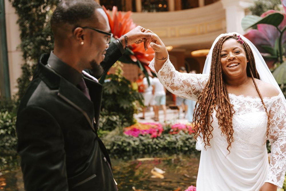 Bride in white lace dress with long braids laughs as groom in black suit photographs her at indoor garden wedding.