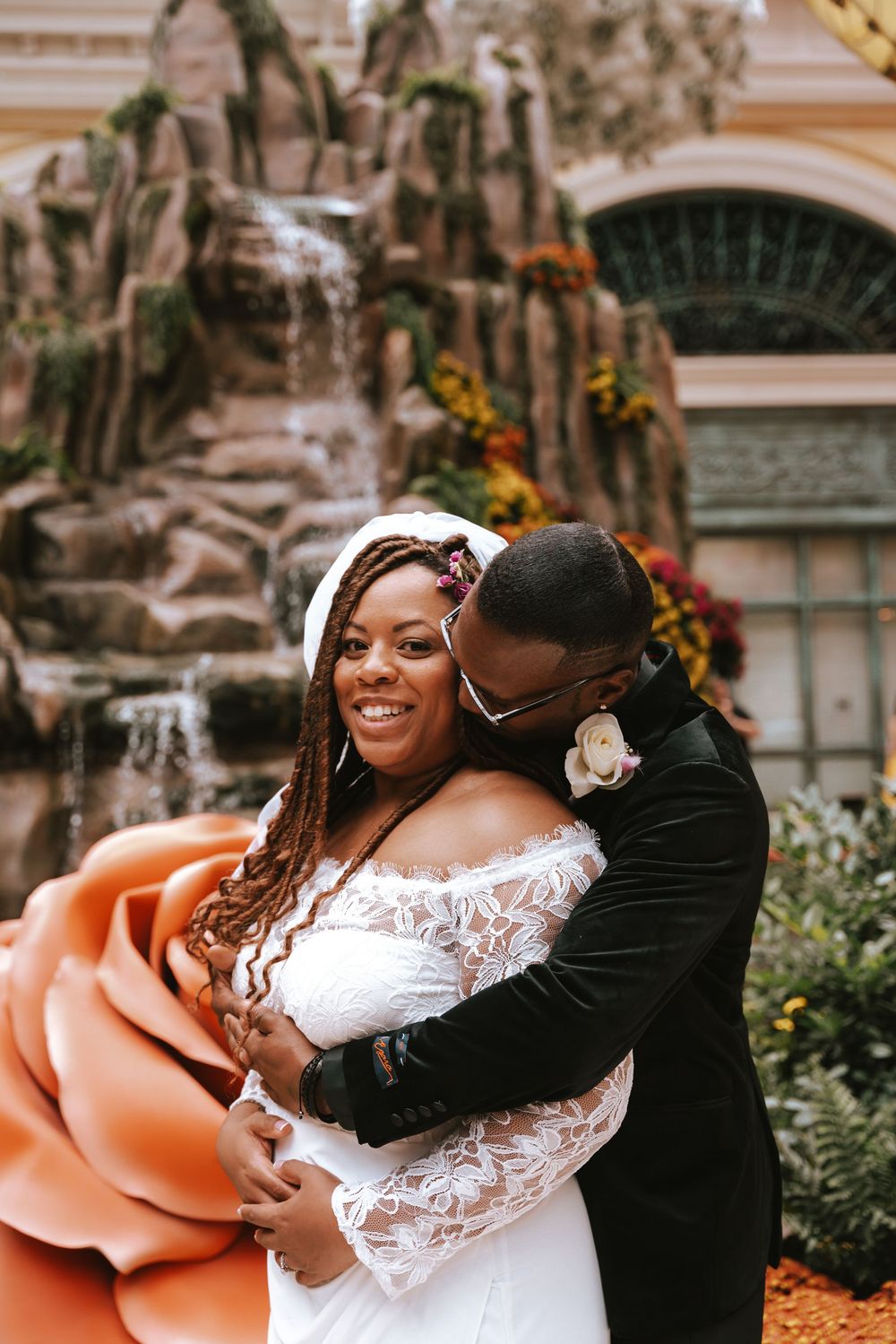 A joyful bride in lace gown embraces groom in black velvet suit near a waterfall at Bellagio Las Vegas.