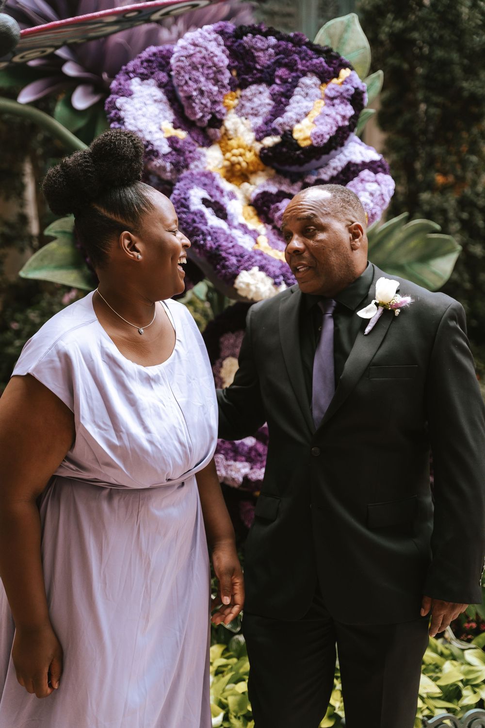 A couple in wedding attire smiles at each other in front of a large purple floral arrangement.