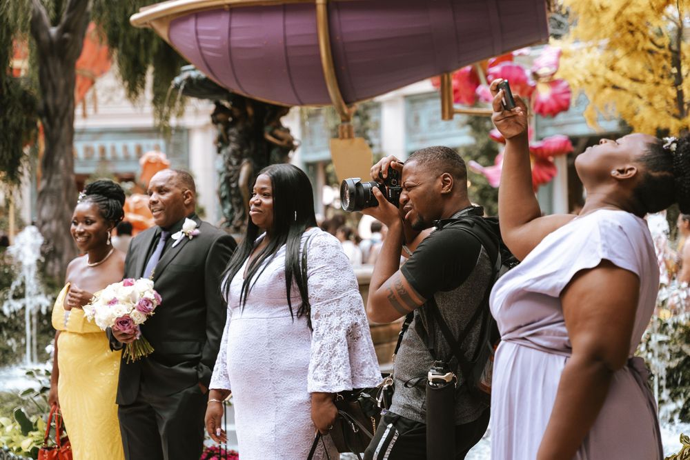Wedding party and photographer capturing moments outdoors, with guests in formal attire including yellow and lavender dresses.