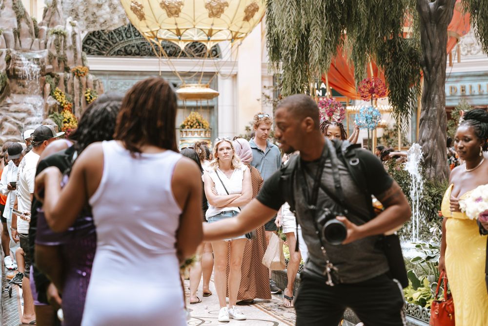 A photographer with a camera greets guests in an ornate indoor venue filled with tourists and tropical decor.