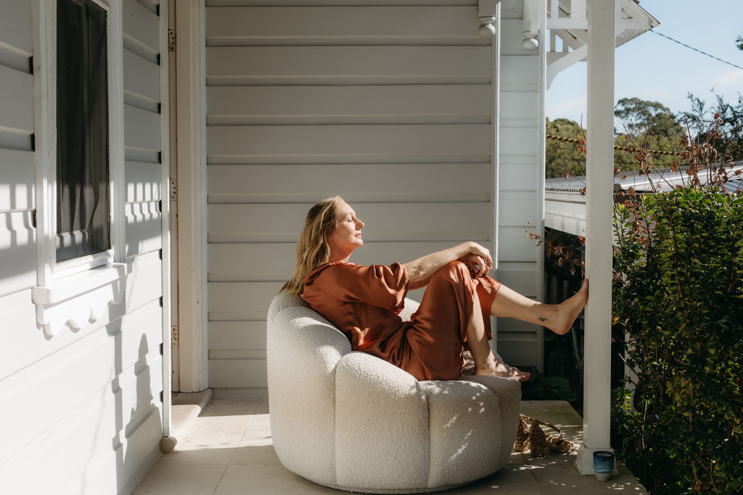 Woman relaxing in a white bean bag chair on a sunny porch wearing a rust-colored outfit.