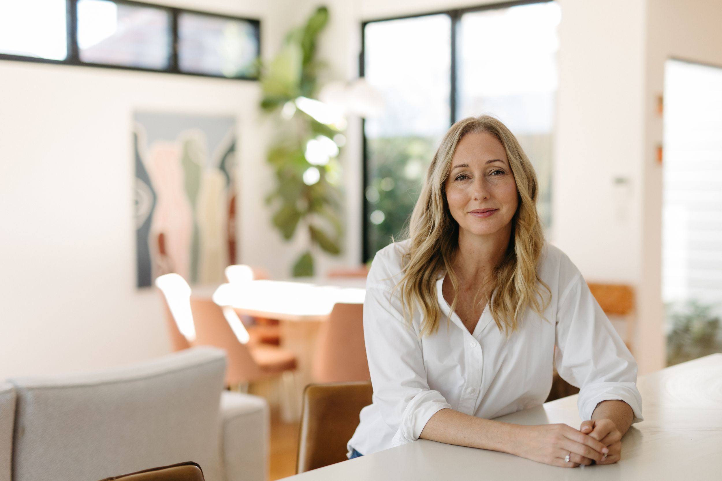 Smiling blonde woman in white shirt sitting at a modern bright home interior table.
