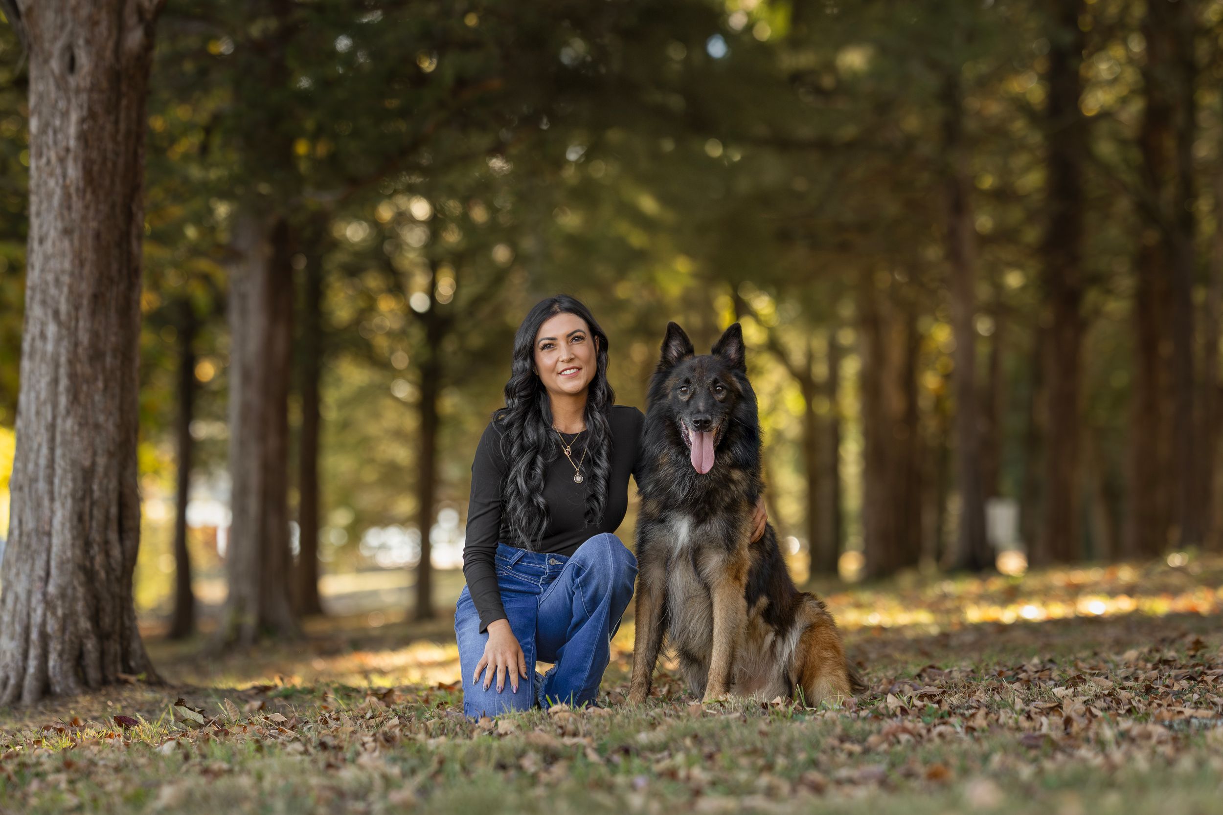 Woman kneeling beside a German Shepherd dog in a tree-lined park with autumn leaves on the ground.