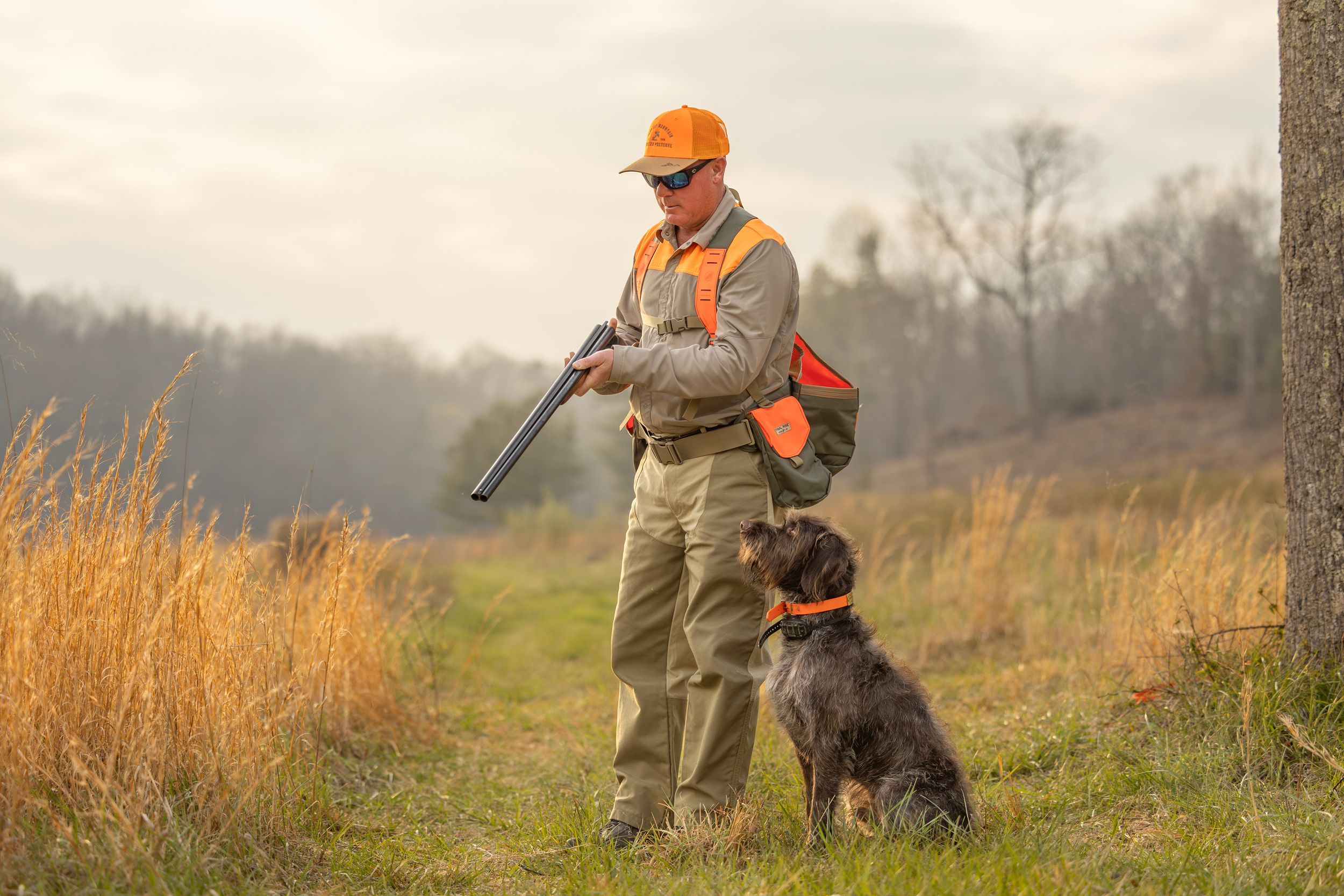Hunter in orange vest and cap stands with shotgun beside a sitting dog on a grassy trail in autumn woods.