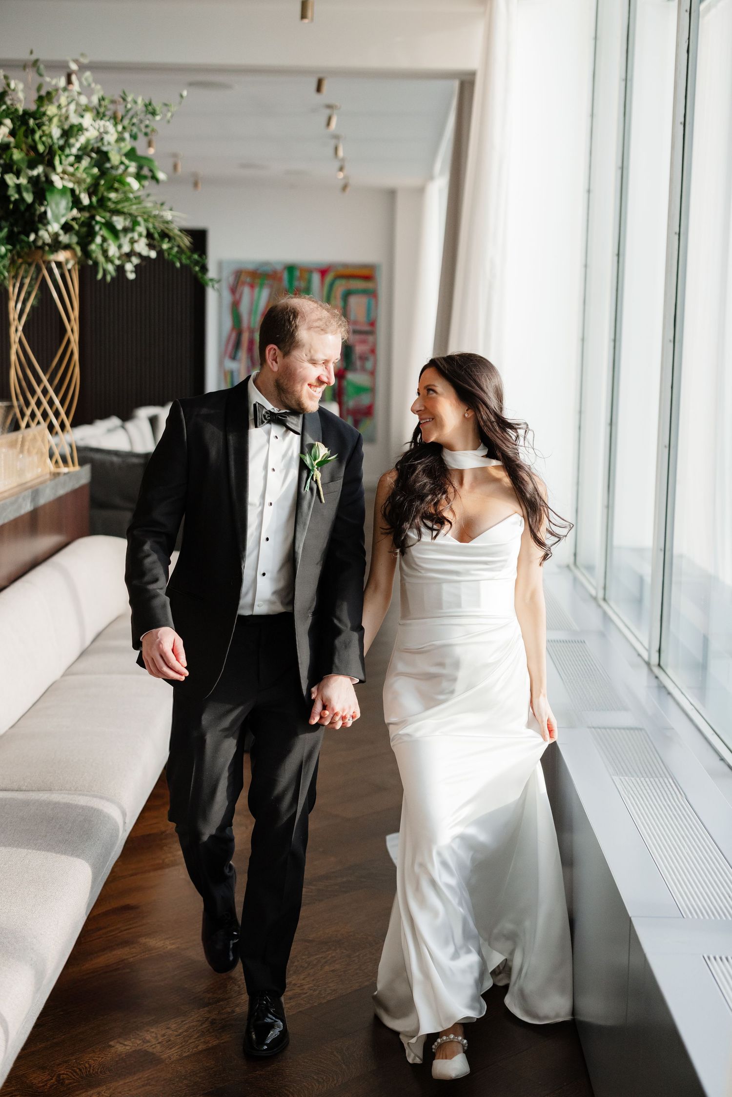 A bride and groom walk hand in hand, smiling at each other, in a modern venue with floor-to-ceiling windows.