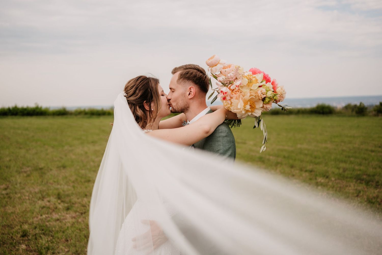 Bride and groom share a romantic kiss in a green field, with wedding veil flowing and colorful bouquet held high.