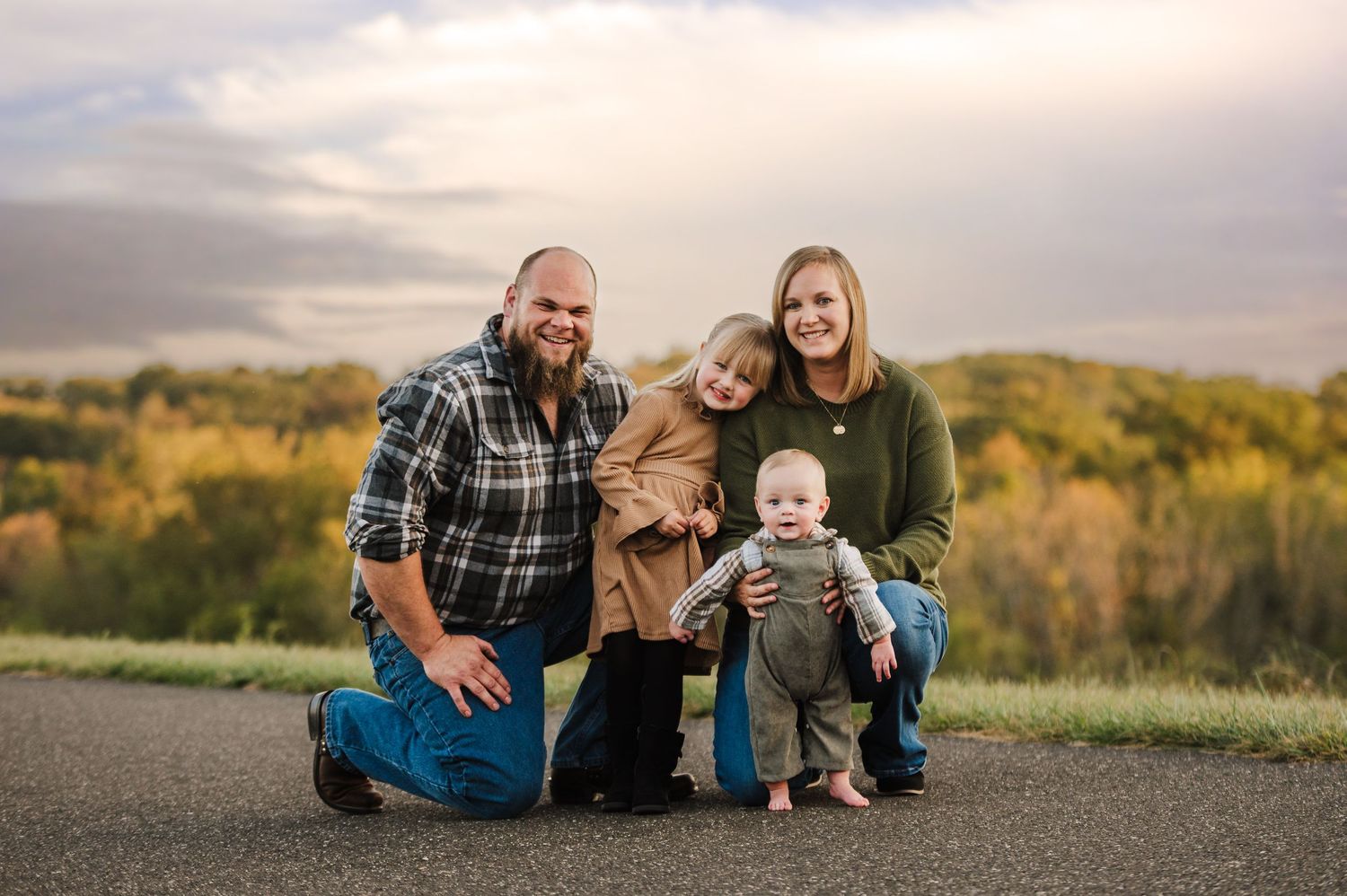Family of four posing outdoors in autumn, parents kneeling with two young children, colorful fall foliage in background.