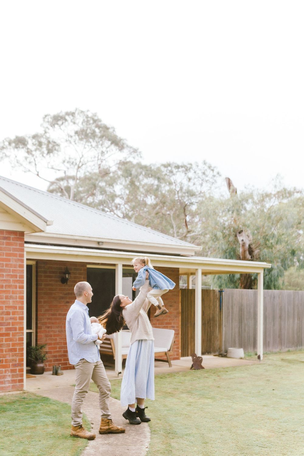 Family playing in backyard, mother lifting child while father watches outside brick home.