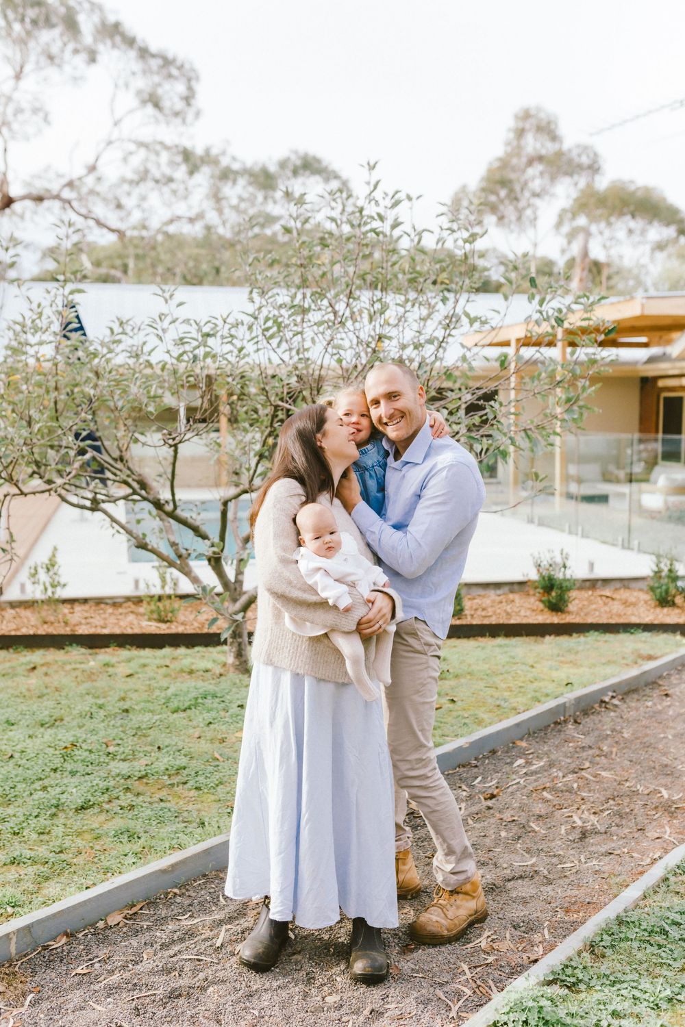 Happy family posing outdoors, woman holding baby while kissing mans cheek near olive tree.