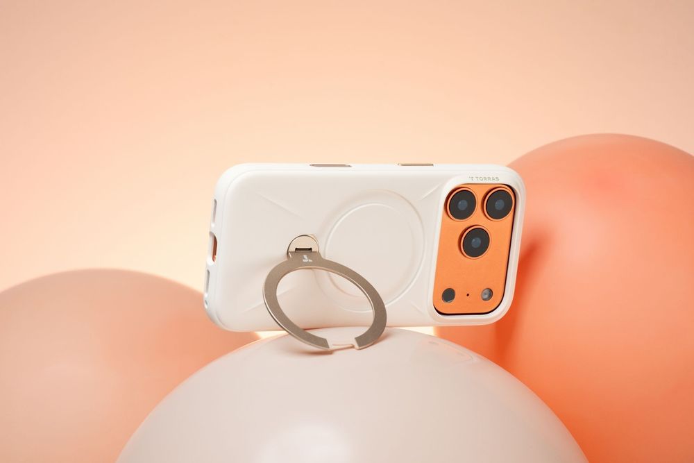 White phone case with ring holder and MagSafe compatibility, displayed among peach and pink balloons on a warm background.