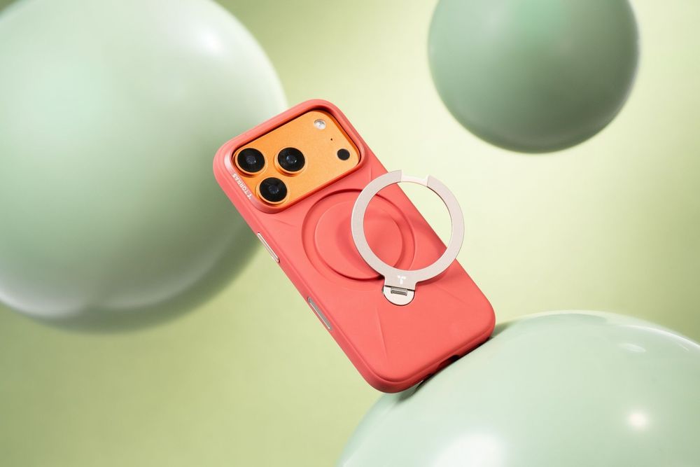 A coral pink iPhone case with a MagSafe ring holder floating among sage green spheres on a light green background.