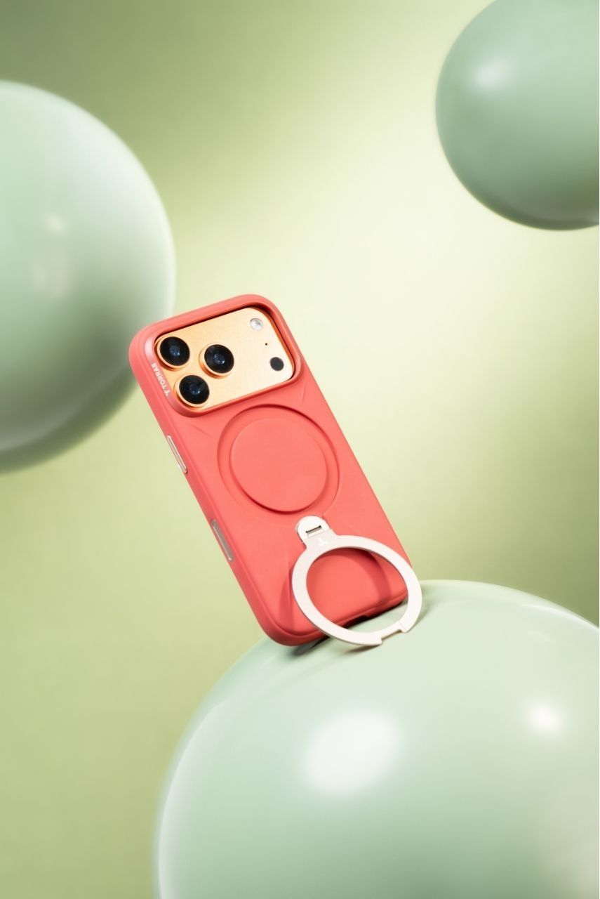 Coral red iPhone case with white ring stand resting on sage green balloon in matching green setting.