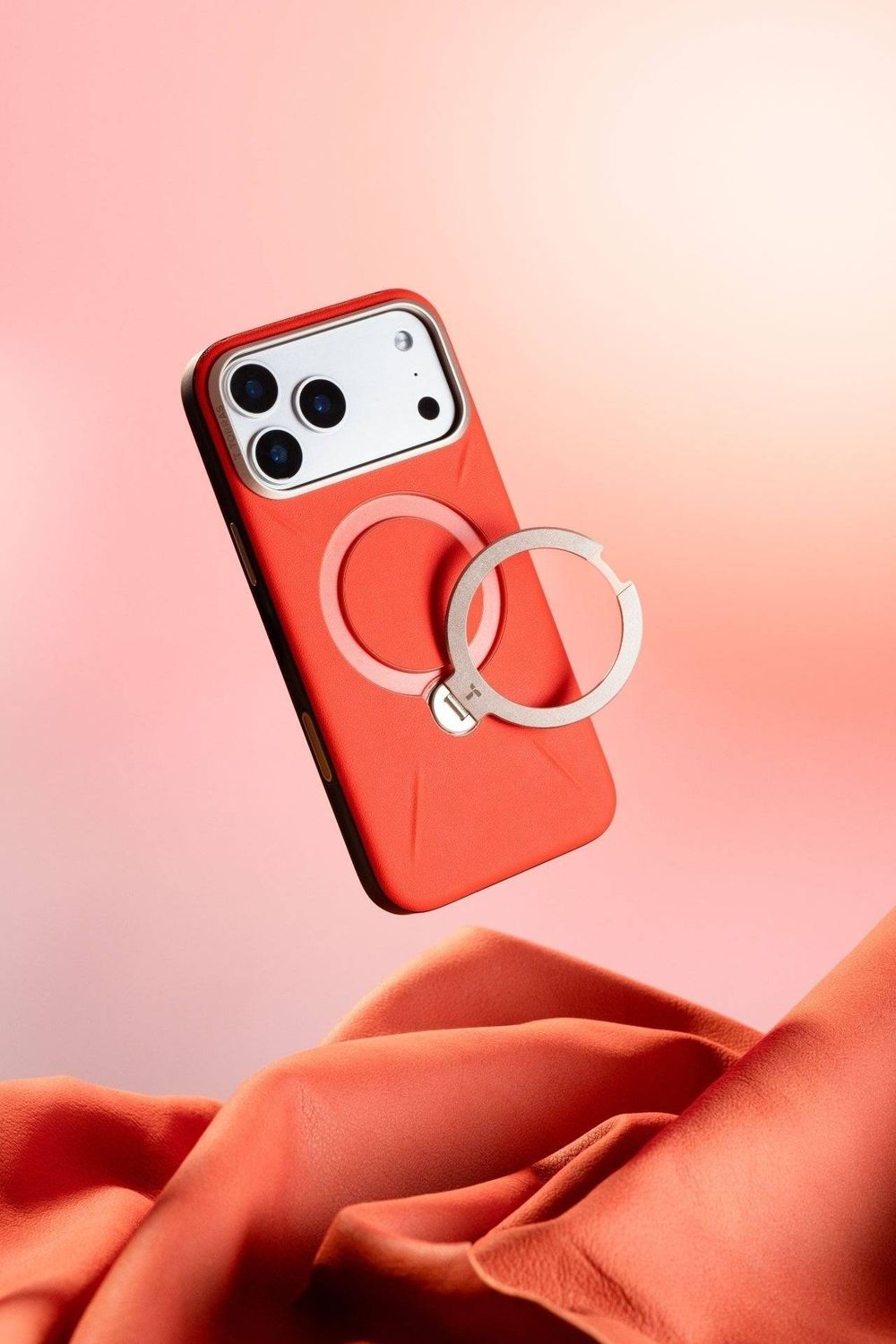 Red iPhone case with MagSafe ring holder floating above coral fabric on pink background.