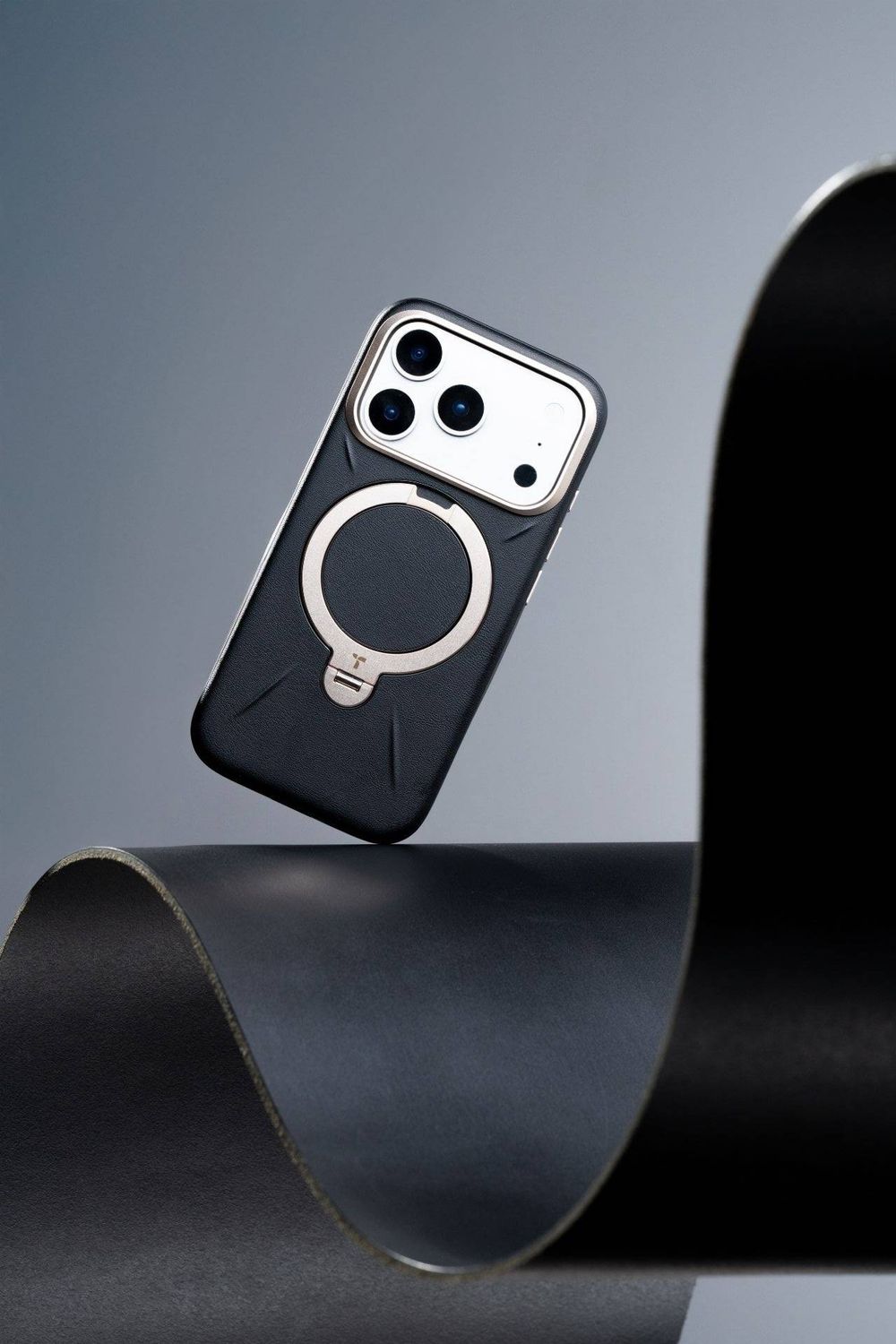 Black MagSafe iPhone case with silver ring stand, balanced on curved dark paper against gray background.