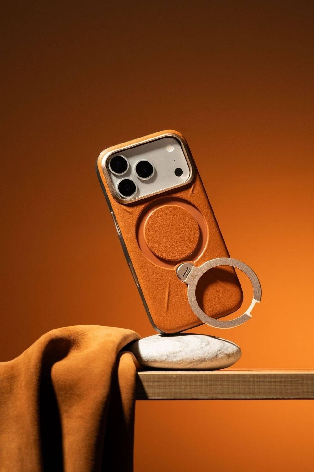 Orange iPhone case with MagSafe ring holder displayed on marble stand against warm orange background.