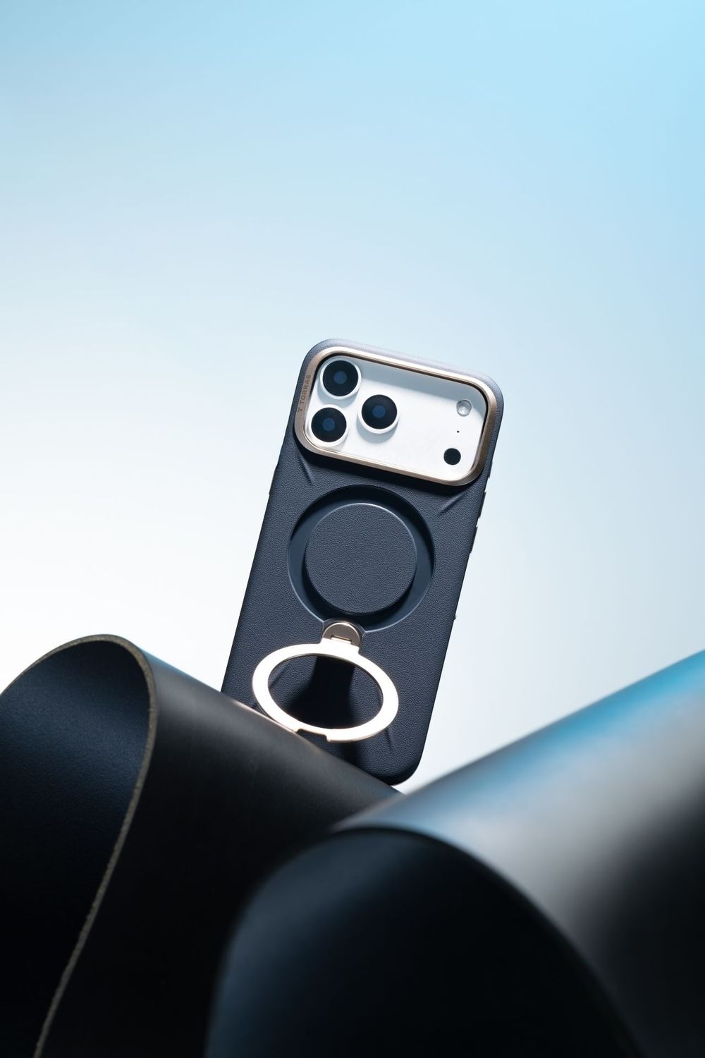 Black iPhone case with MagSafe ring holder displayed upright between curved dark surfaces against a light blue background.