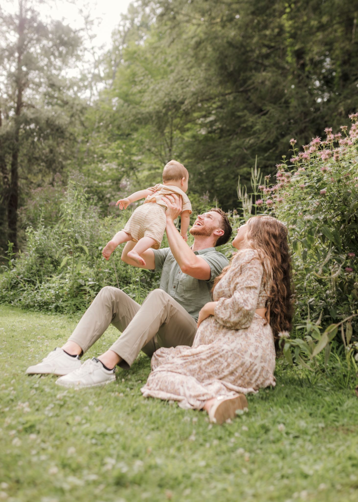 A joyful family sits on grass in a lush garden, father lifting baby while mother watches and smiles warmly.