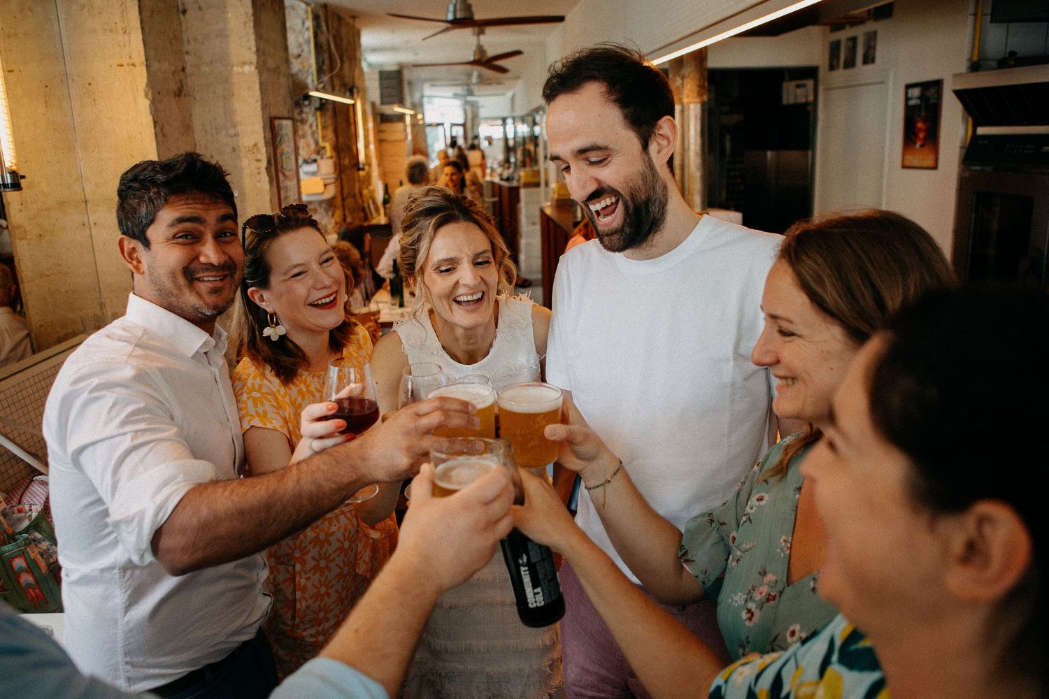 Group of friends laughing and toasting with beer and wine glasses at a casual indoor gathering.