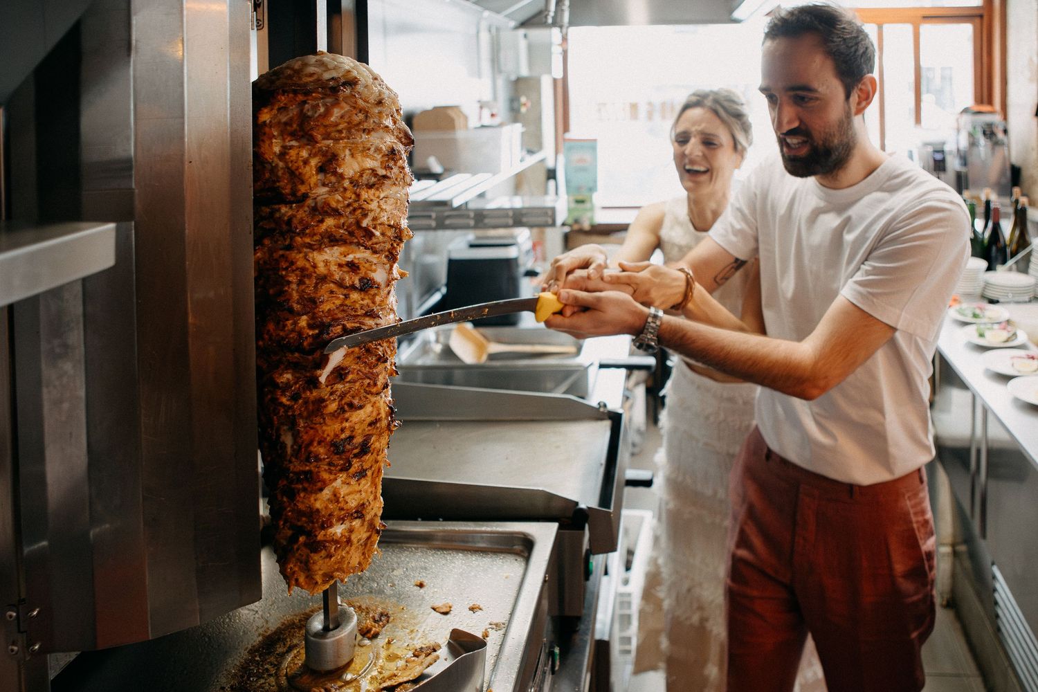 Chef slicing meat from a rotating shawarma spit in a professional kitchen with a female colleague nearby.