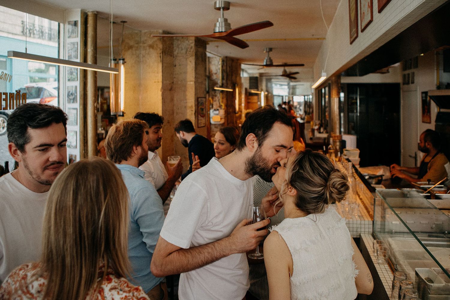 Couple sharing a kiss at a lively bar gathering with guests socializing in a warmly lit restaurant.