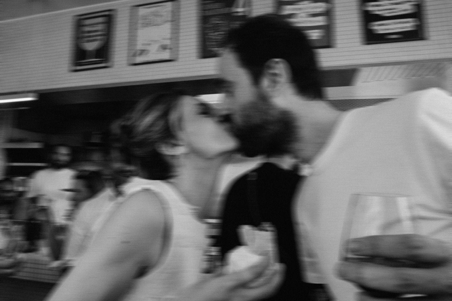 Black and white photo of a couple sharing a romantic kiss at a bar, with wine glasses visible.