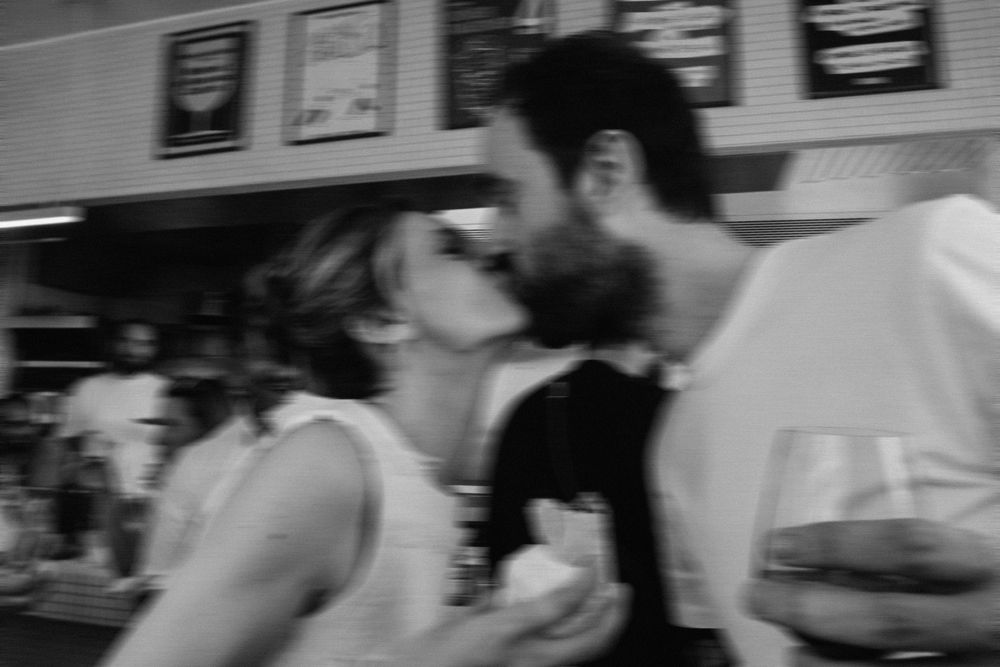 Black and white photo of a couple sharing a romantic kiss at a bar, with wine glasses visible.