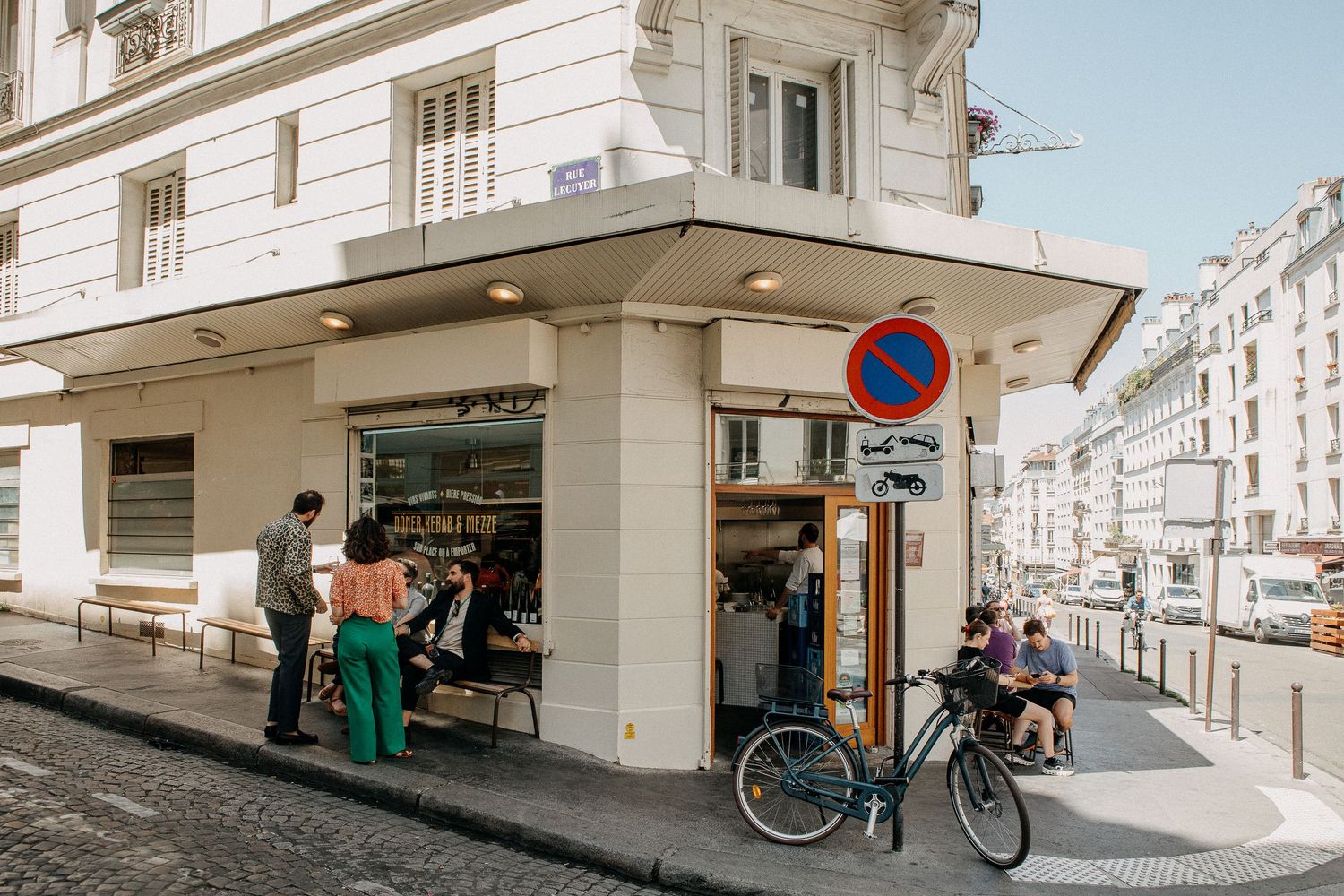 Corner cafe in Paris with patrons browsing outside, a bicycle parked nearby, and a no-parking sign on a sunny day.