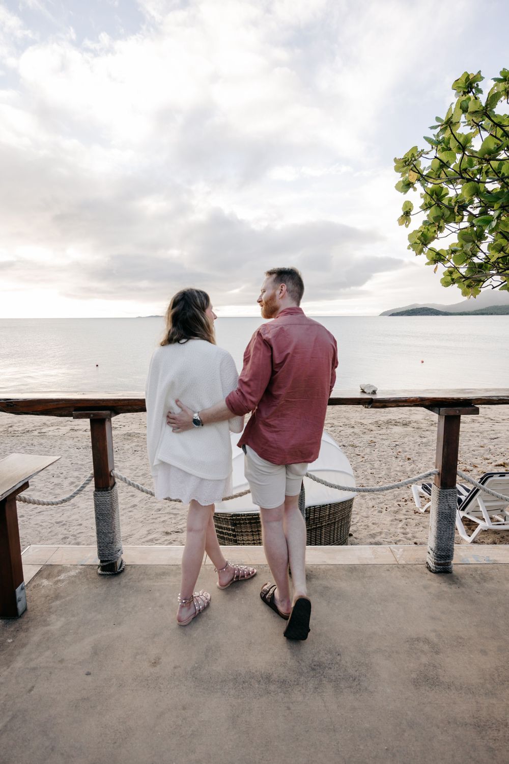 Couple embracing while walking along a tropical beach terrace at sunset, enjoying a romantic seaside view.