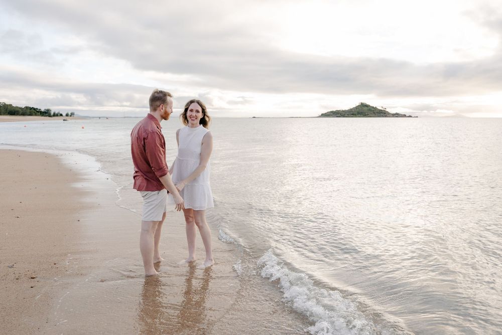 Couple holding hands and walking along a sandy tropical beach at sunset with a small island in the background.