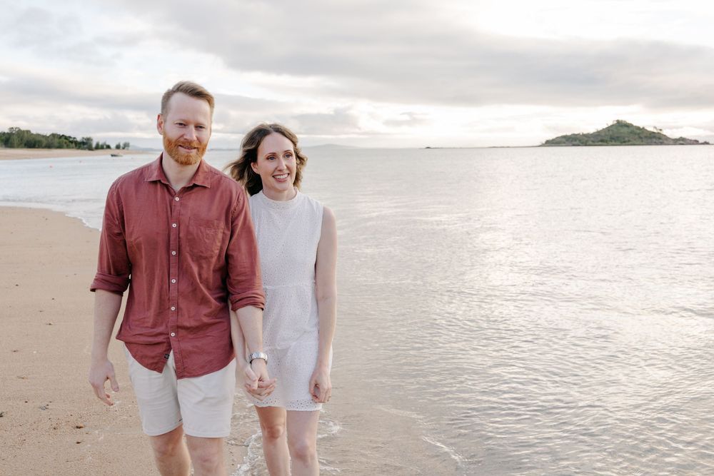 Couple holding hands smiling on a tropical beach at sunset with calm ocean and island in background.