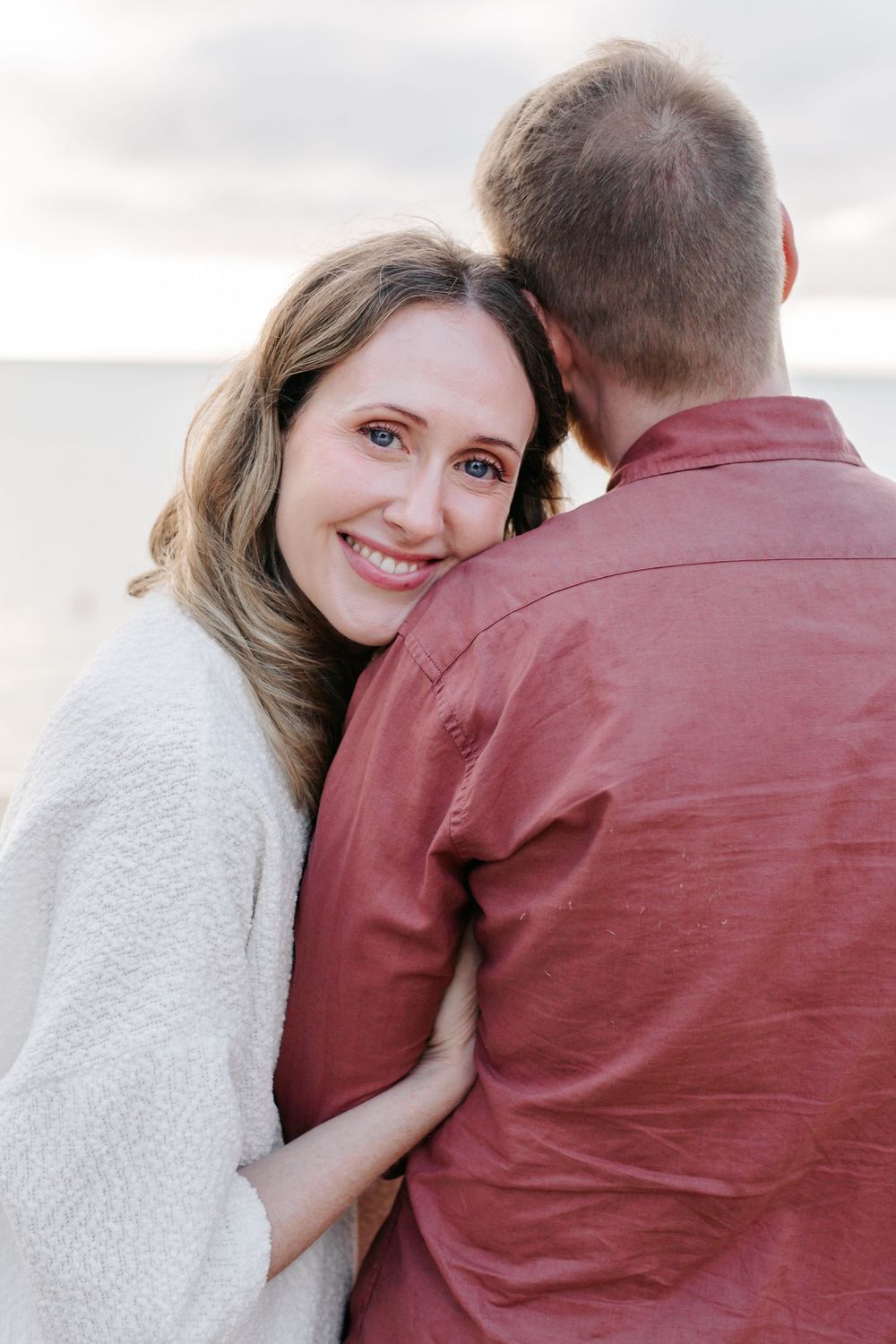 A smiling woman in a white sweater embraces a man in a red shirt on a beach, resting her head on his shoulder.