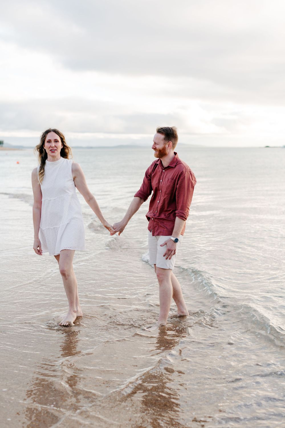 A couple holds hands walking barefoot on a sandy beach, woman in white dress and man in red shirt, with ocean waves.