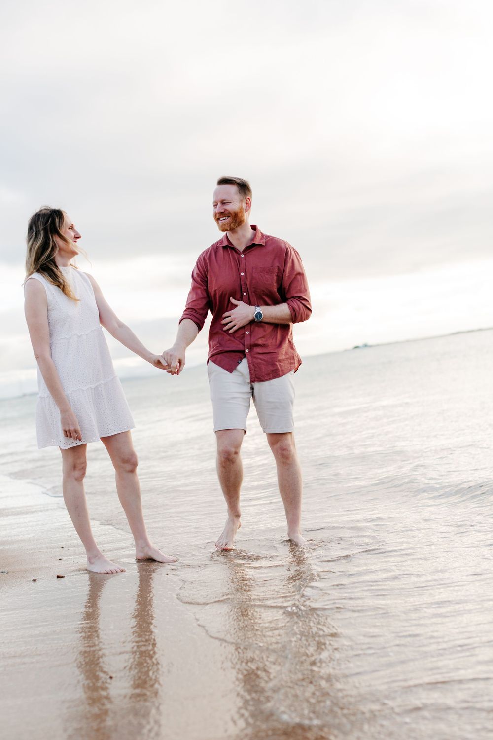 A happy couple holds hands while walking barefoot on a sandy beach, smiling at each other on a cloudy day.