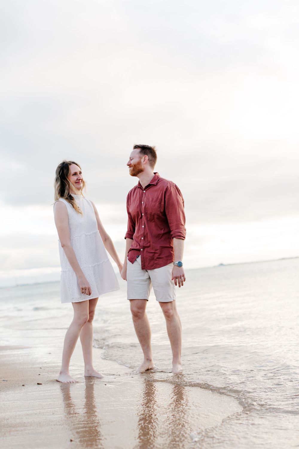 Couple holding hands and smiling at each other on a sandy beach, woman in white dress, man in red shirt and shorts.