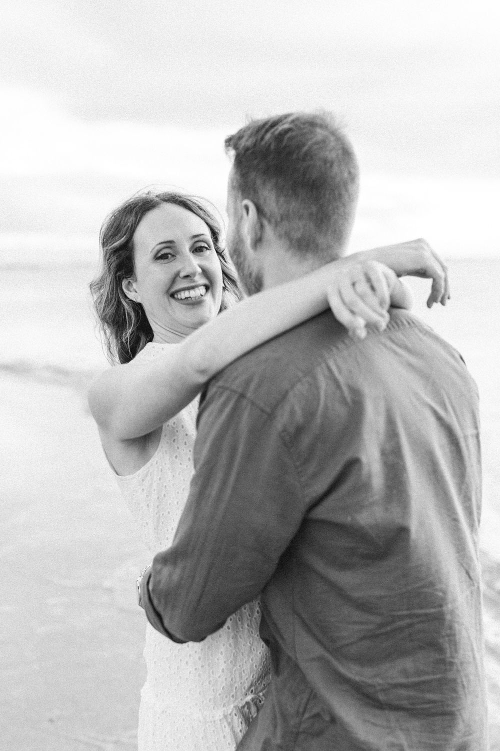 Black and white photo of a smiling woman embracing a man on a beach, capturing a joyful romantic moment.