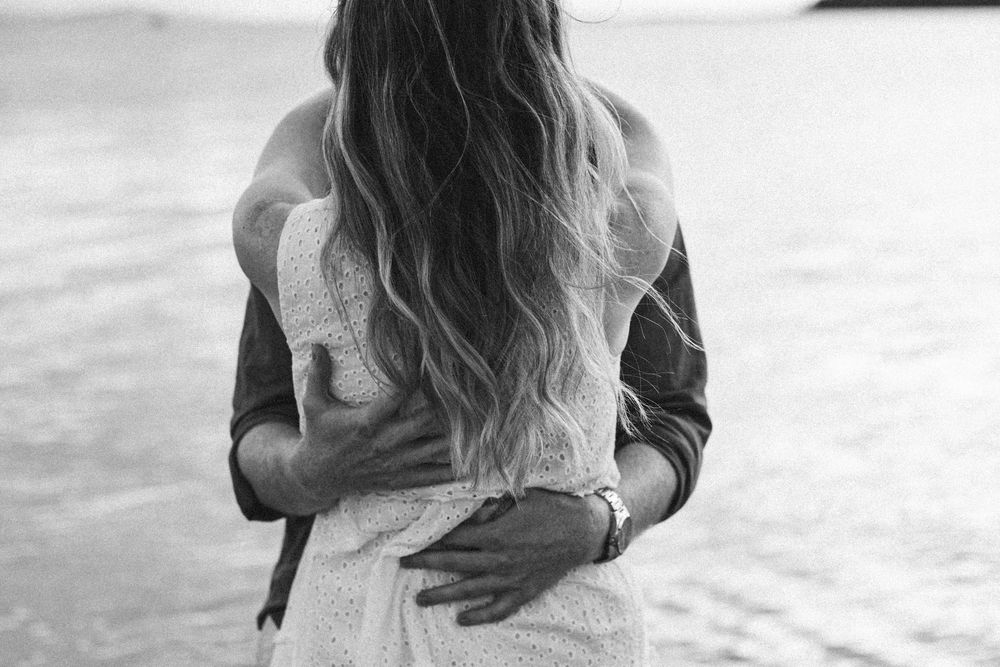 Black and white photo of a couple embracing near the ocean, woman with long wavy hair in white dress.
