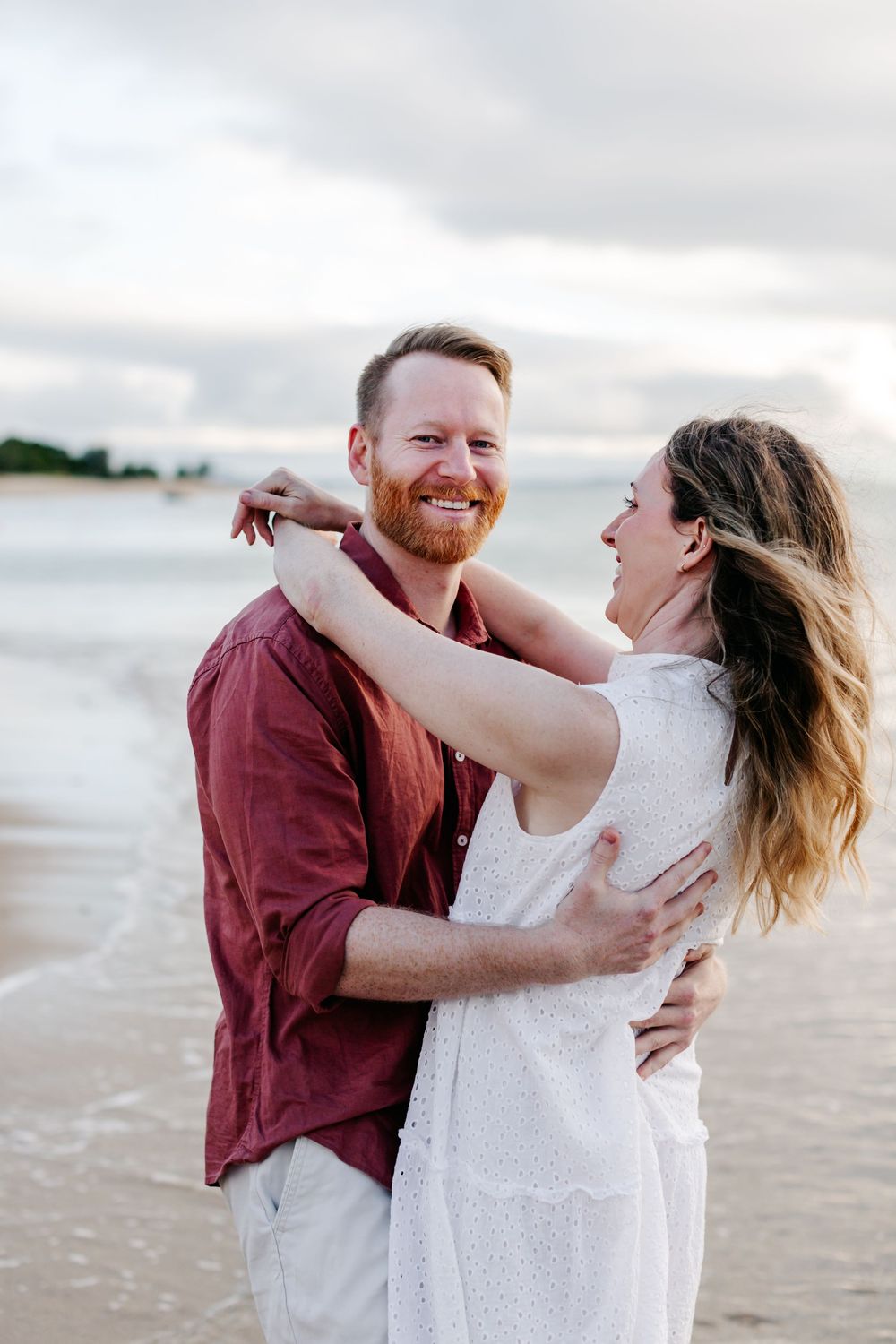 A happy couple embraces on a beach at sunset, man in red shirt and woman in white dress smiling.
