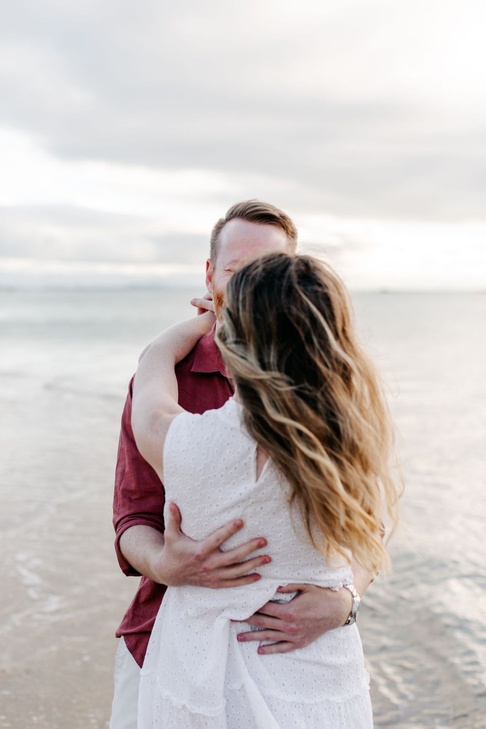 Couple embracing and kissing on a beach with ocean waves and cloudy sky in the background.