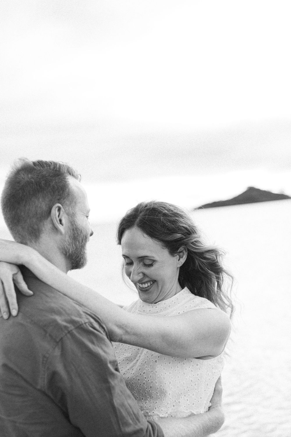 Black and white photo of a smiling couple embracing on a sandy beach with a mountain in the background.