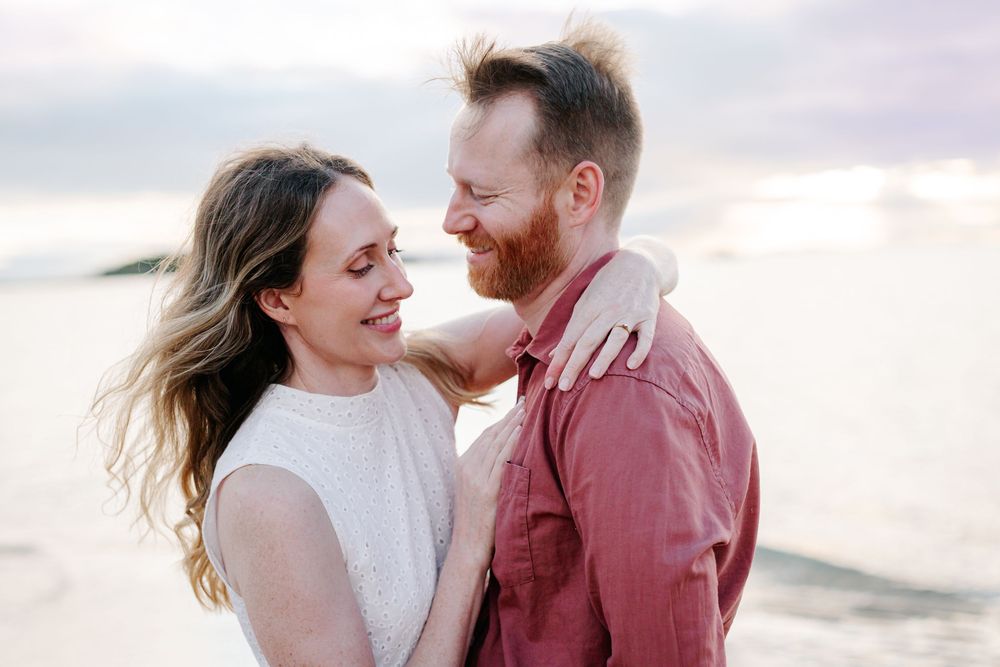 Couple smiling at each other on a beach at sunset, woman in white dress, man in red shirt.