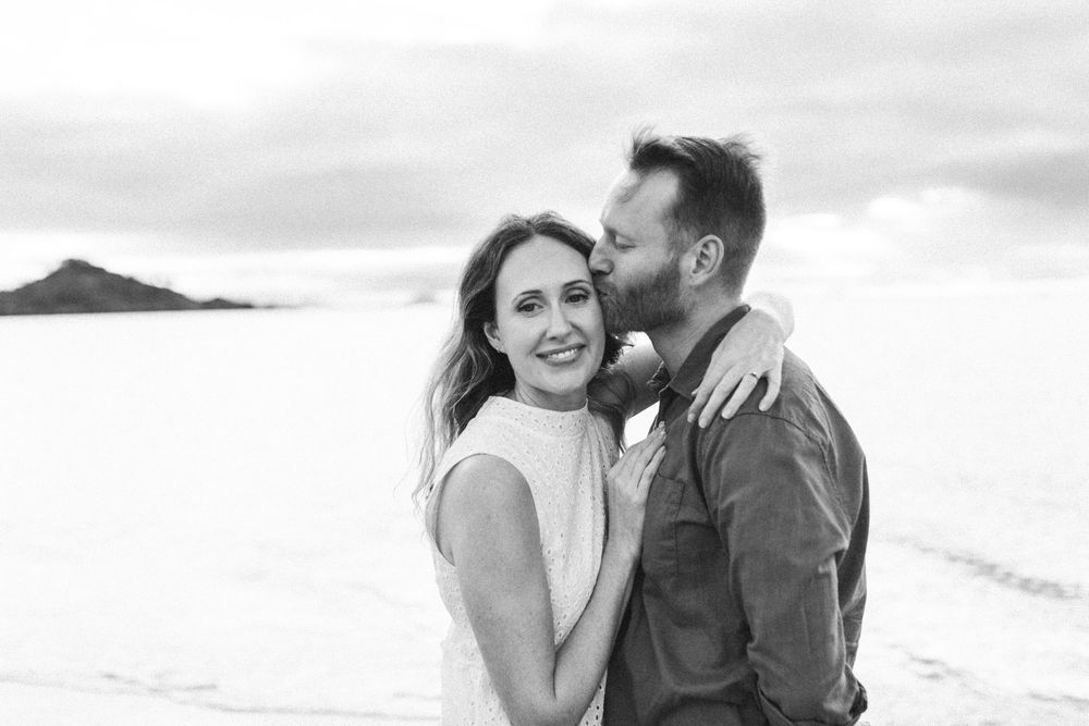 Black and white photo of a smiling couple embracing outdoors, man kissing woman's cheek near a scenic landscape.